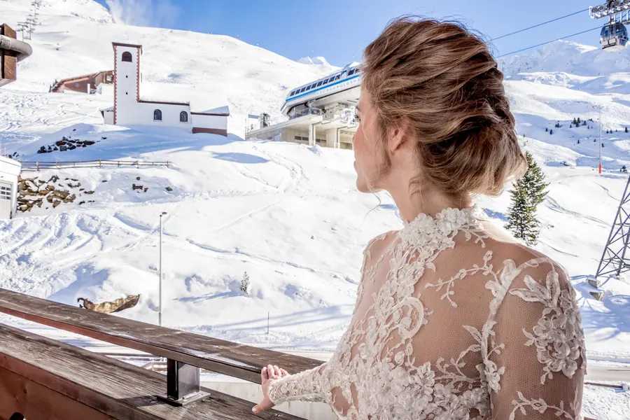 Bride in a lace dress looking out at a snow-covered mountain chapel, winter wedding on the Arlberg